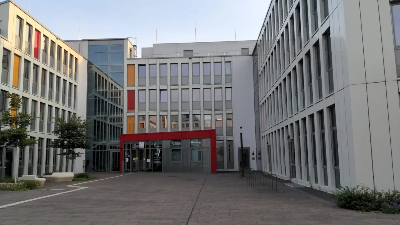Eine Wissenschaftlerin und ein Wissenschaftler arbeiten hinter einer Glasfassade und mischen Chemikalien mit Großgeräten.